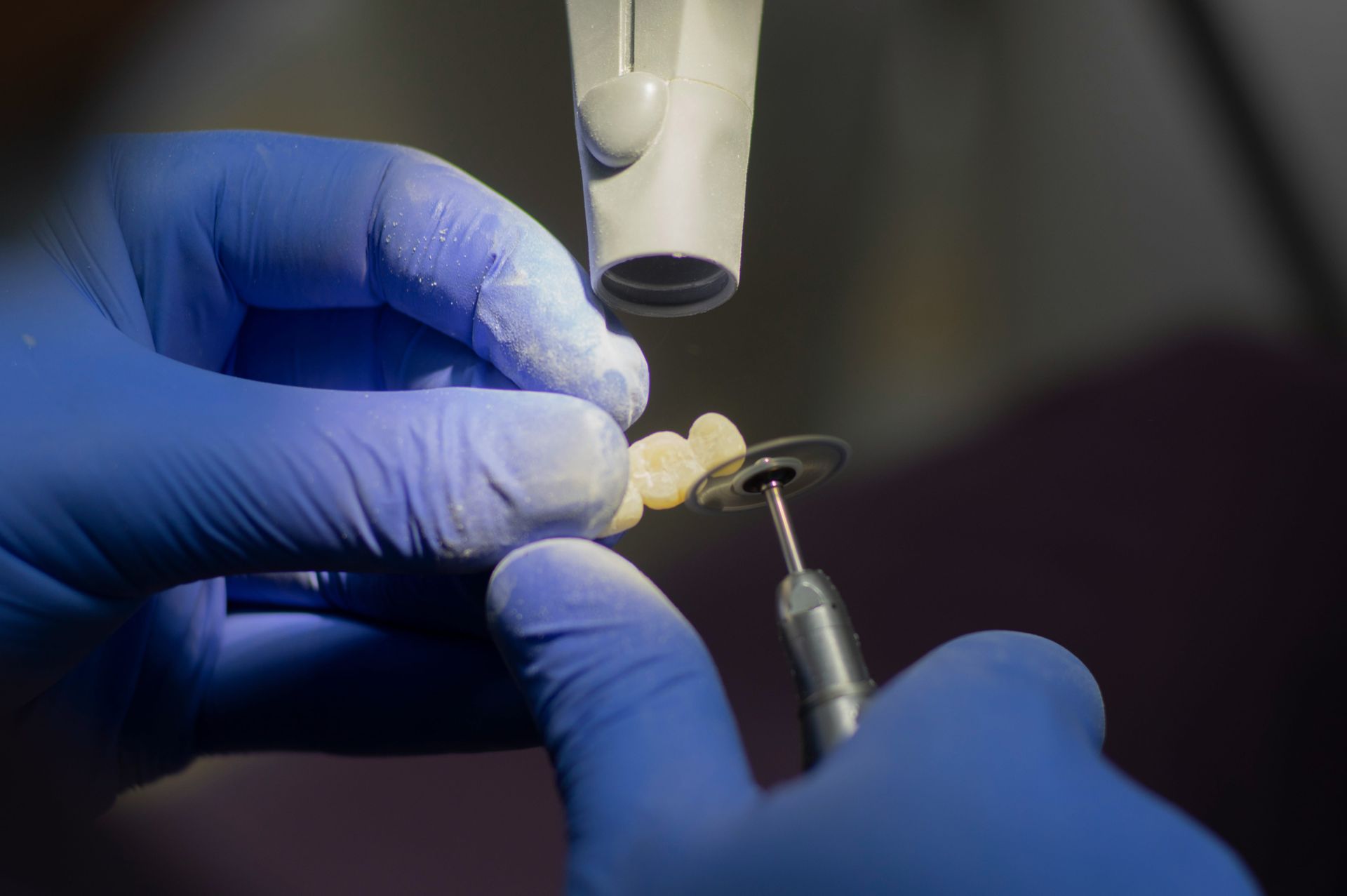 Dentist's gloved hands using a tool to adjust a dental bridge under a bright light.