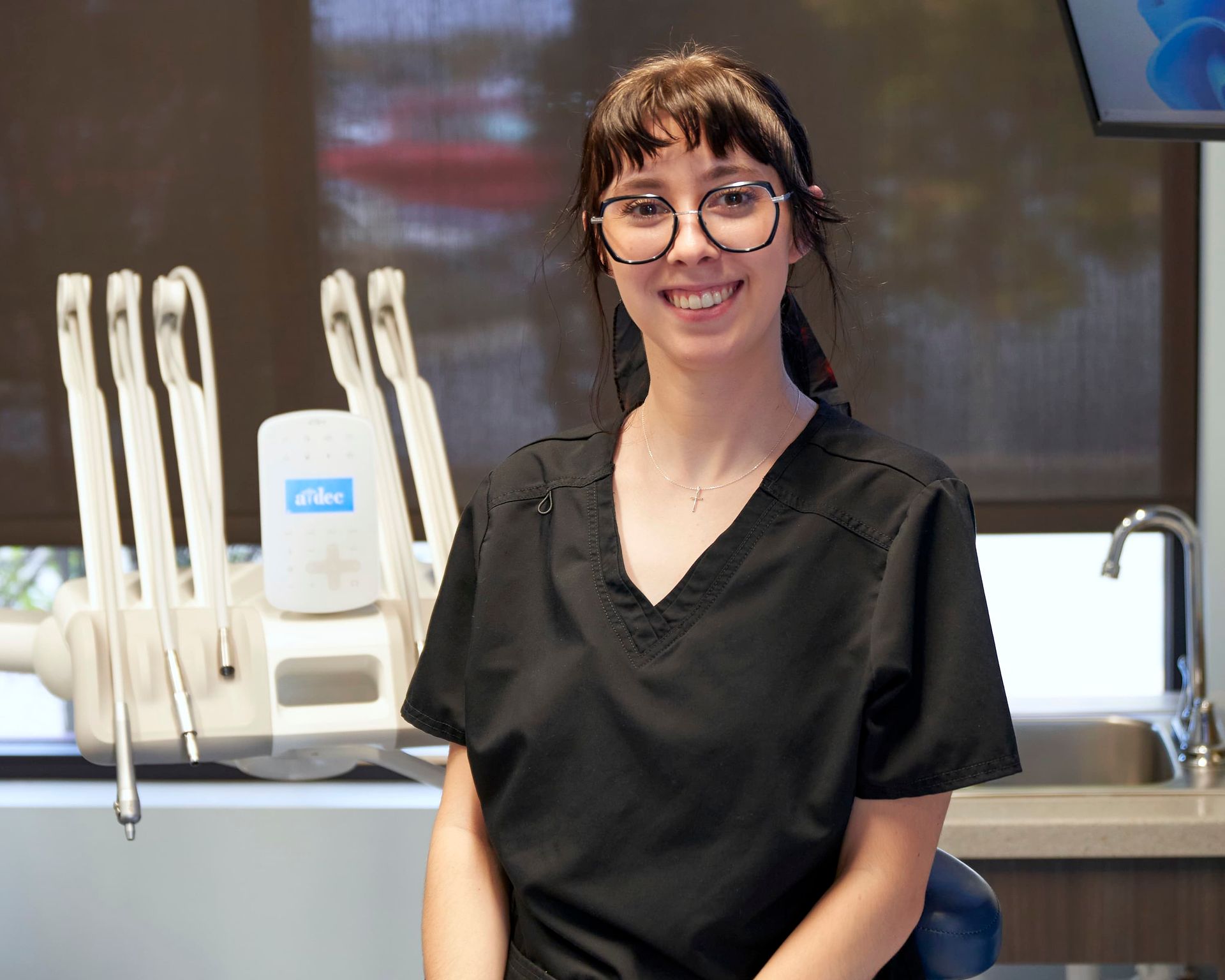 Woman in black scrubs and glasses smiles in a dental office.