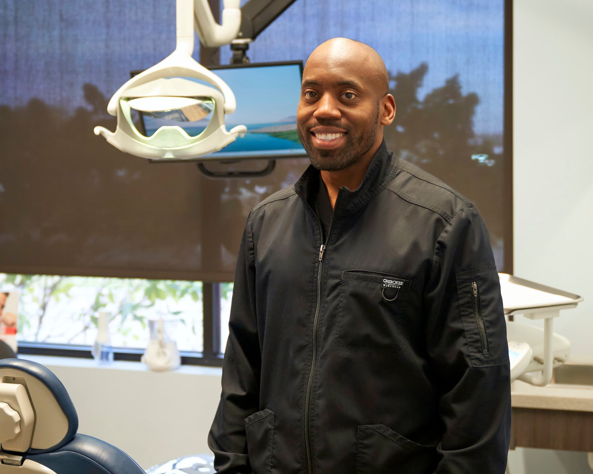 Man in black jacket smiles in a dental office. Dental chair, overhead light, and window visible.