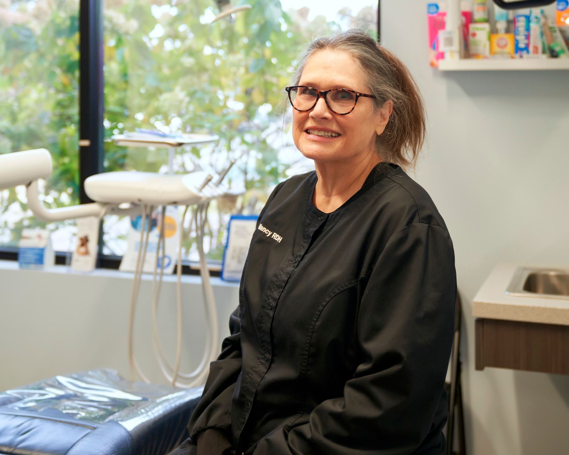 Woman wearing glasses sits in a dental chair. She is in a dental office with a window in the background.