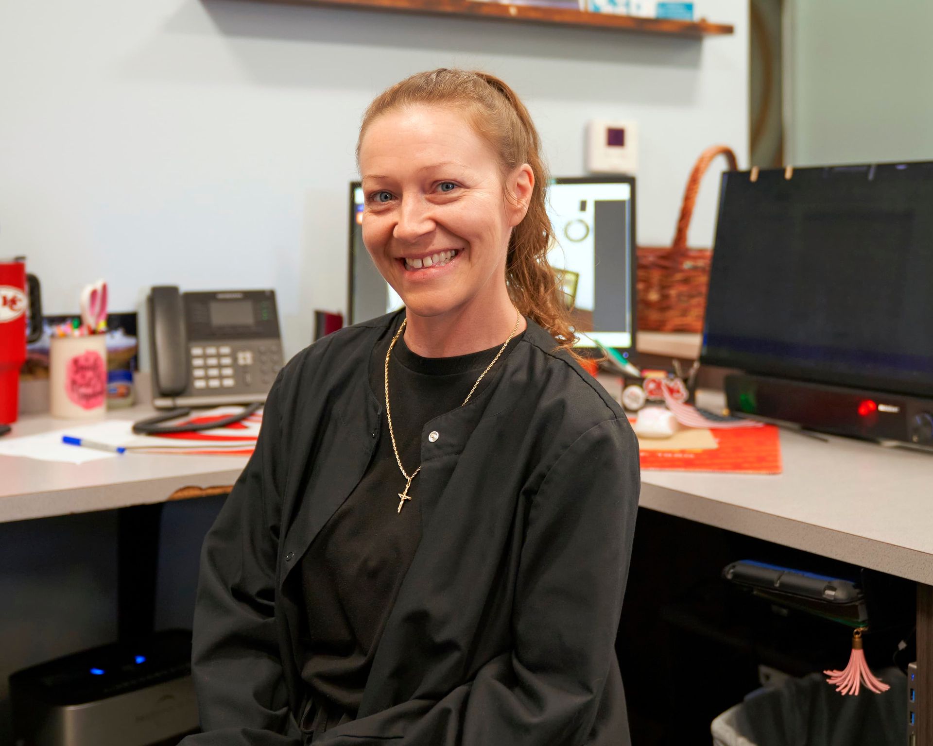Woman with curly brown hair smiling, wearing a dark top, in front of a brick wall.