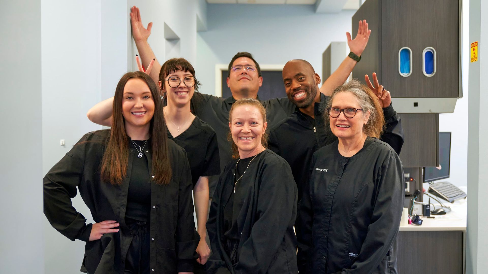 Dental office team smiles for a photo; dark scrubs, arms raised, posing near cabinets and a wall.