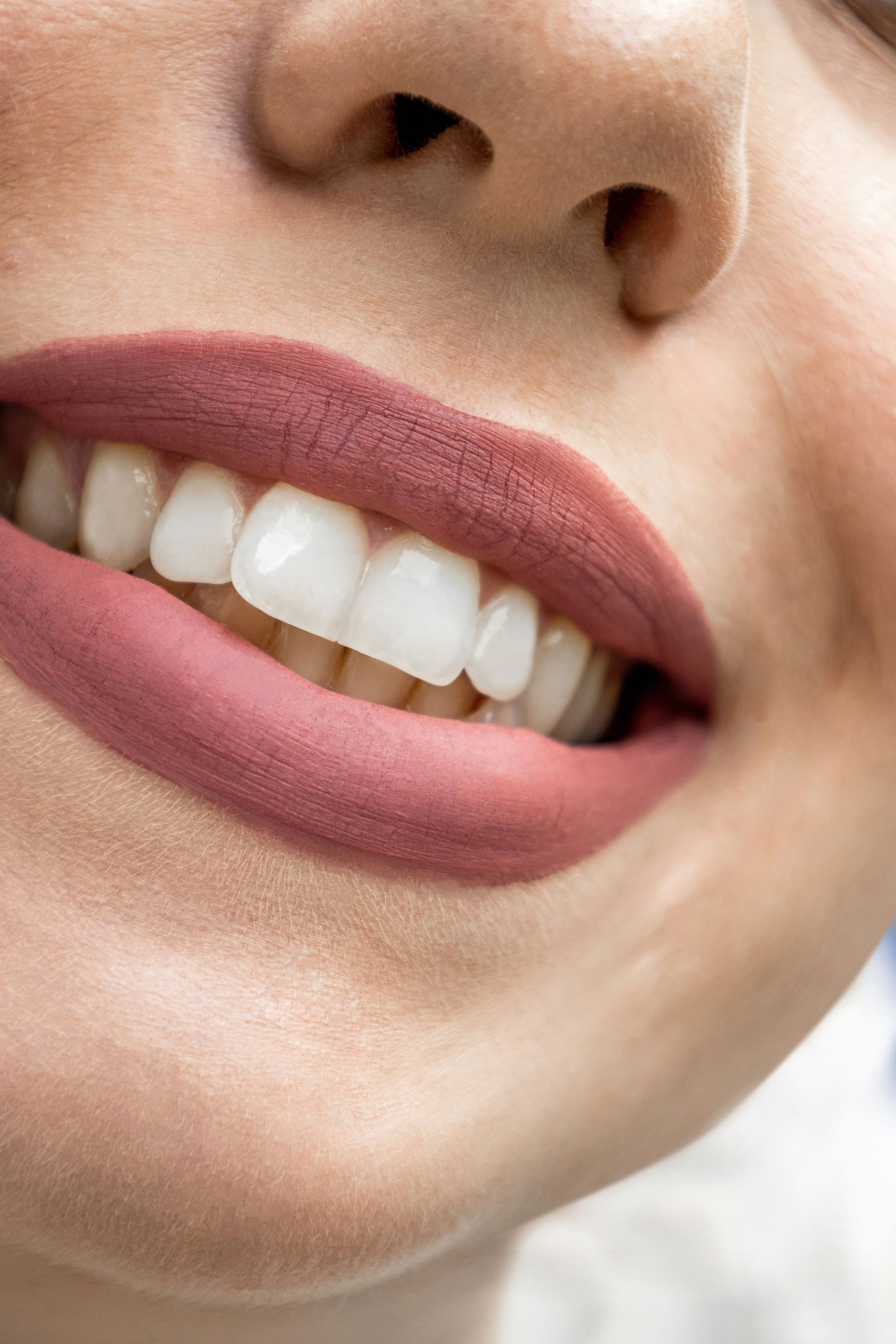 Close-up of a person smiling, showing white teeth and lips with matte pink lipstick.