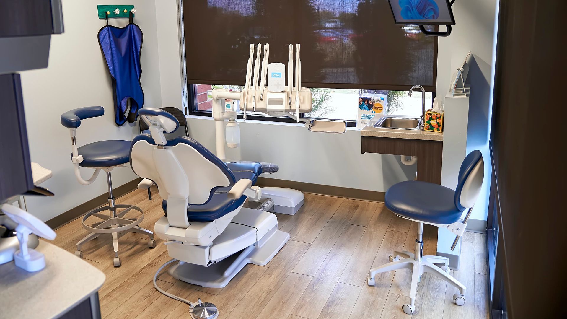 Dental operatory with a chair, tools, and equipment. Light wood floor, dark window blinds, and blue accents.
