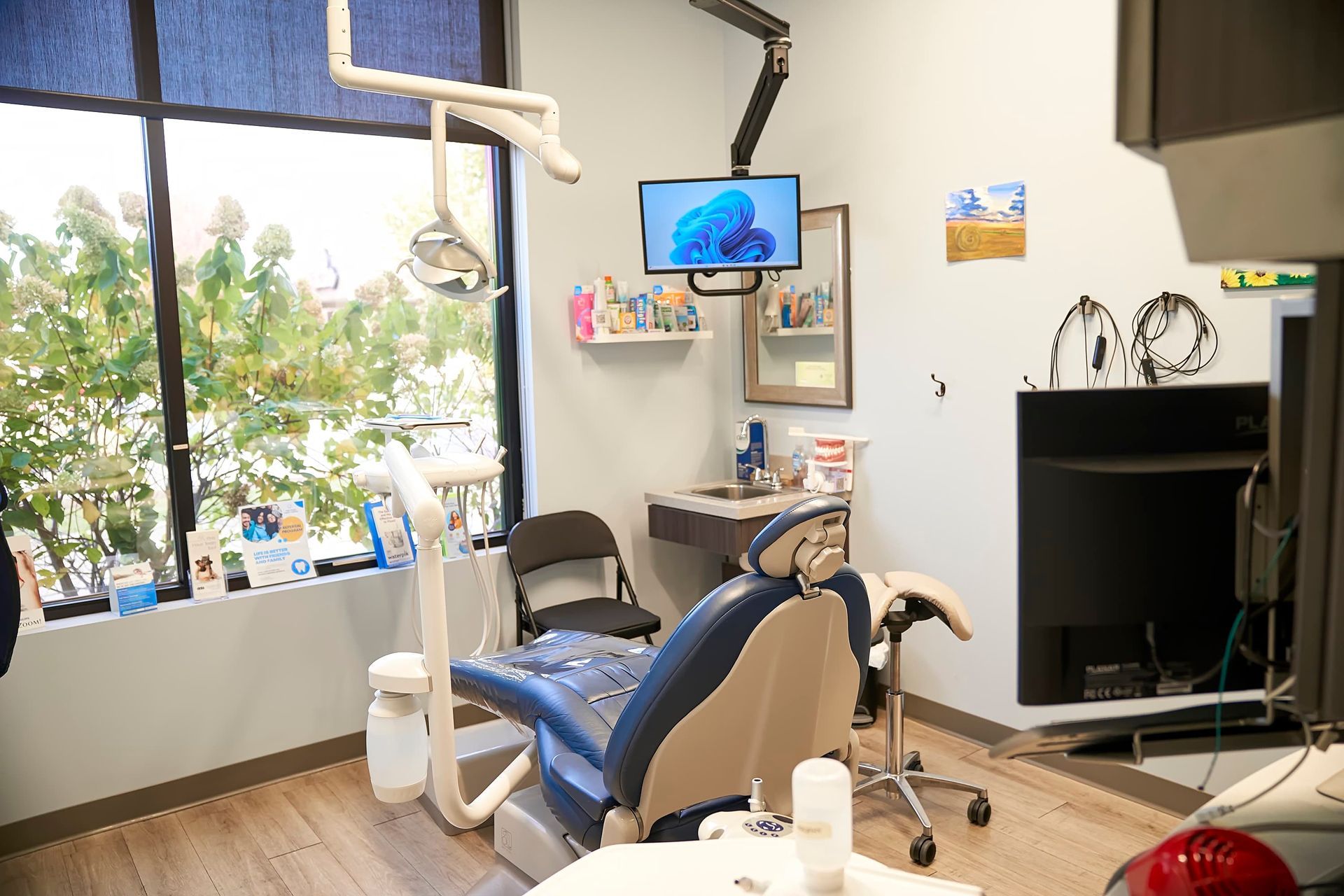 Dental examination room with blue chair, window, monitor, and equipment.