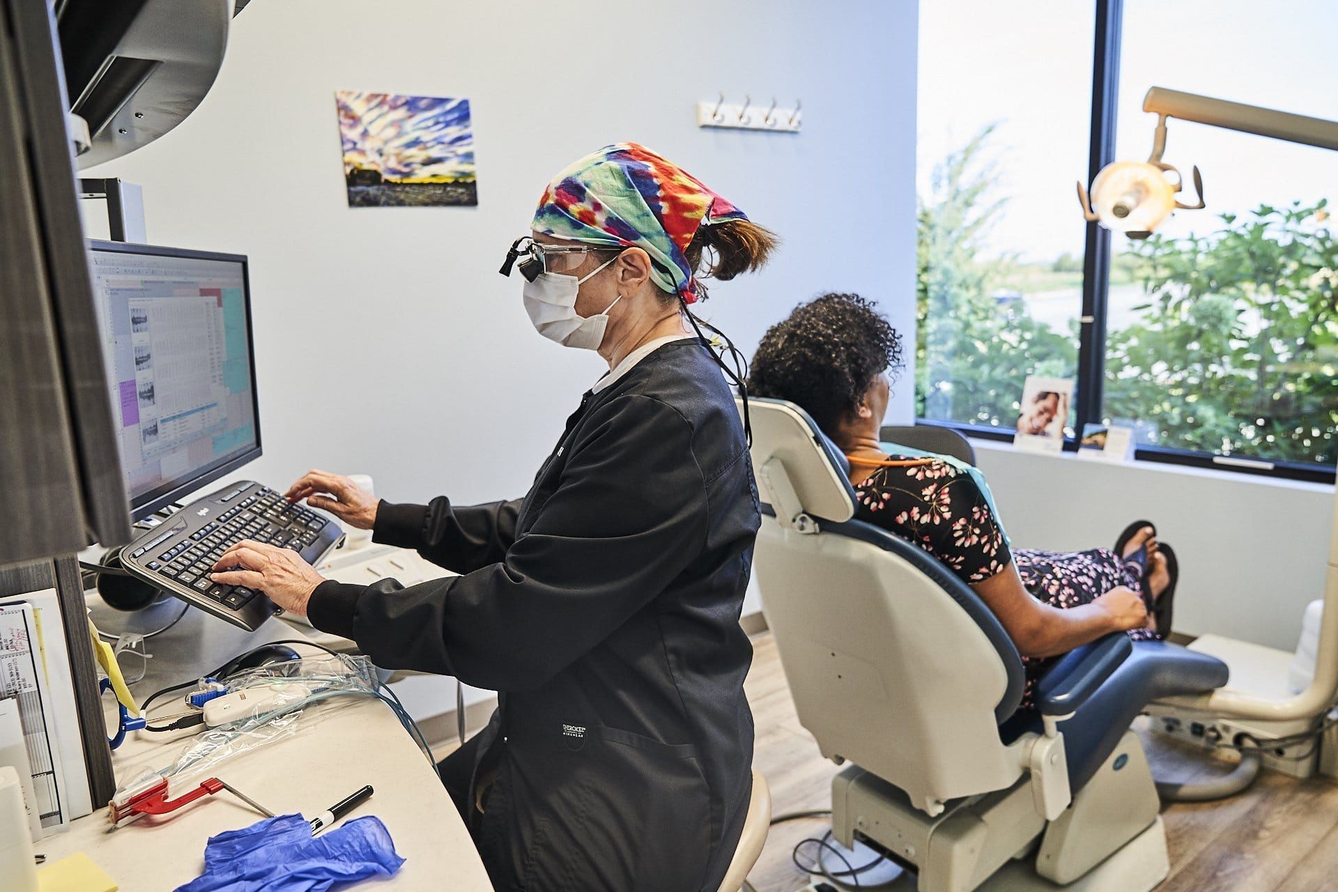A dentist is working on a computer while a patient sits in a dental chair.