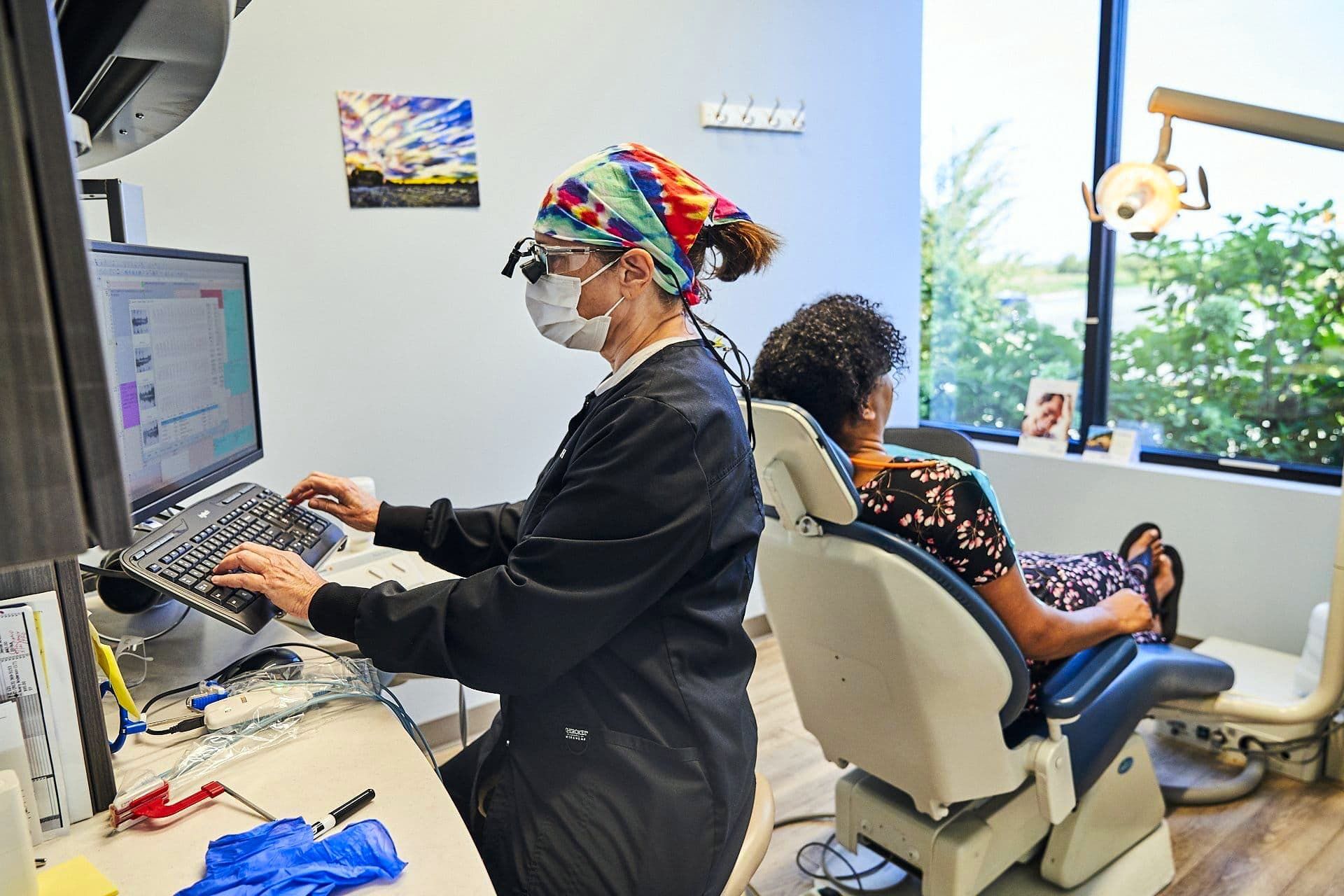 Dentist in mask and cap types on computer. Patient sits in dental chair. Office interior with window.
