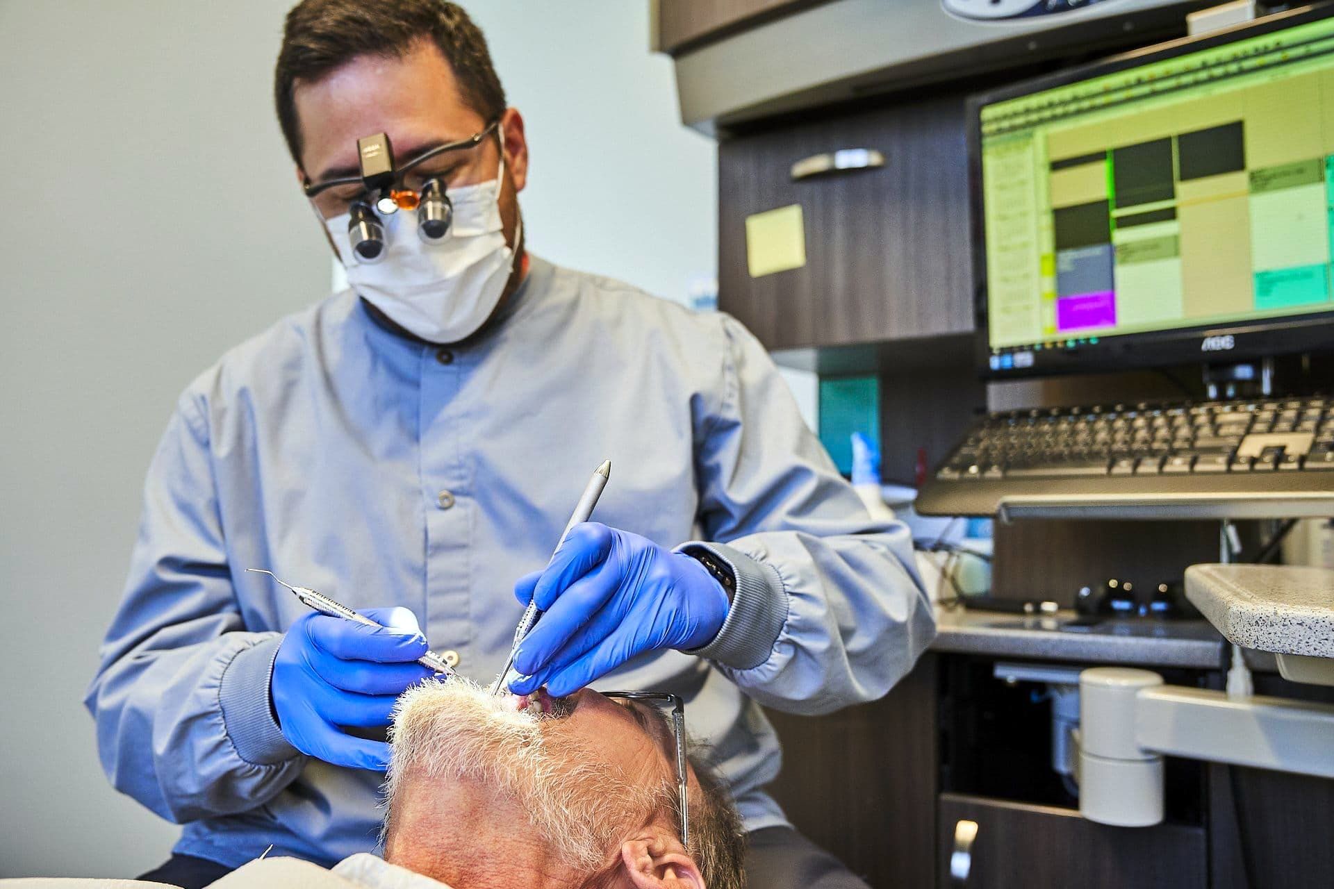 Dentist in mask and loupes, performing procedure on patient's head, dental office setting.