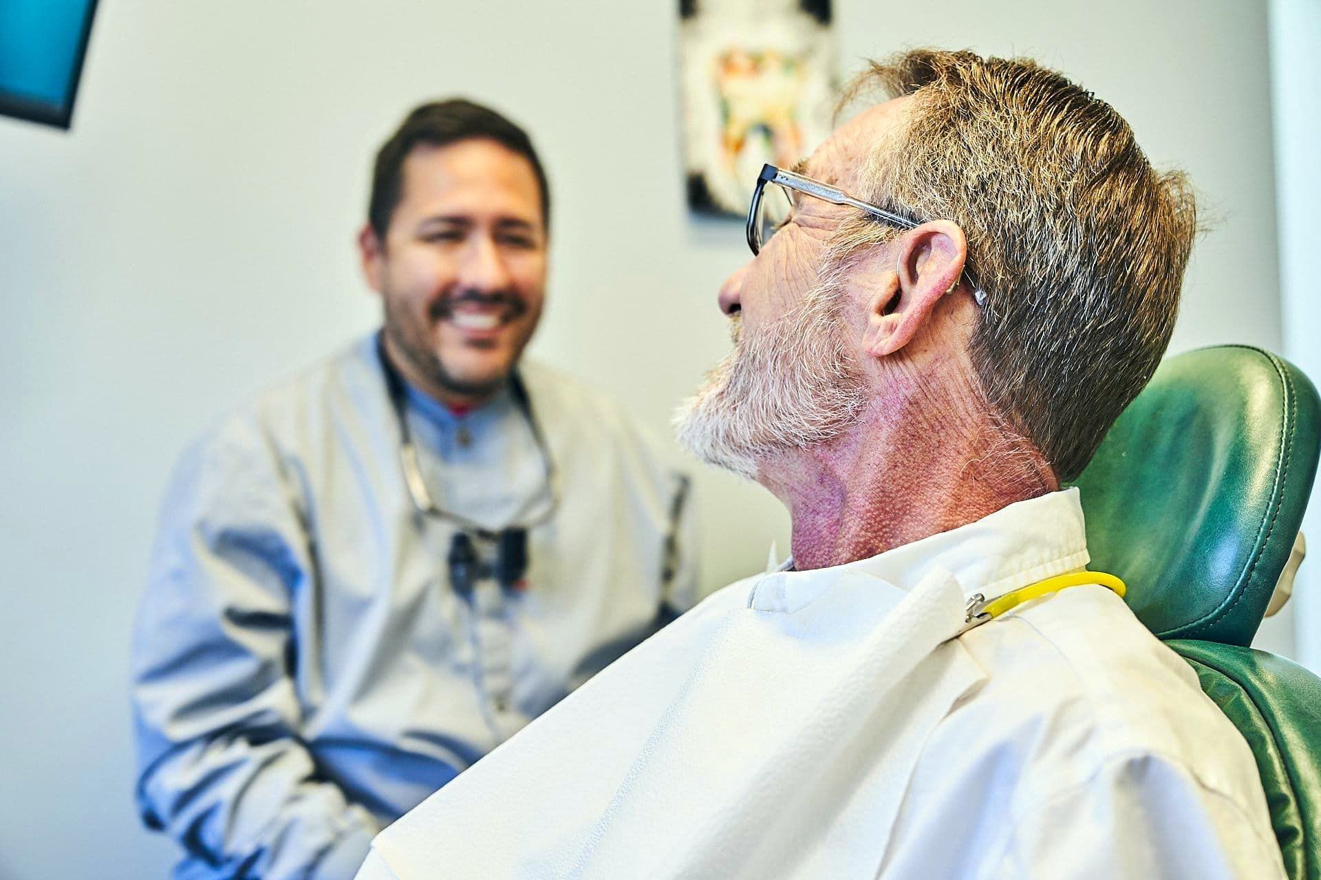 Dentist smiles at patient in dental chair, light-filled office.