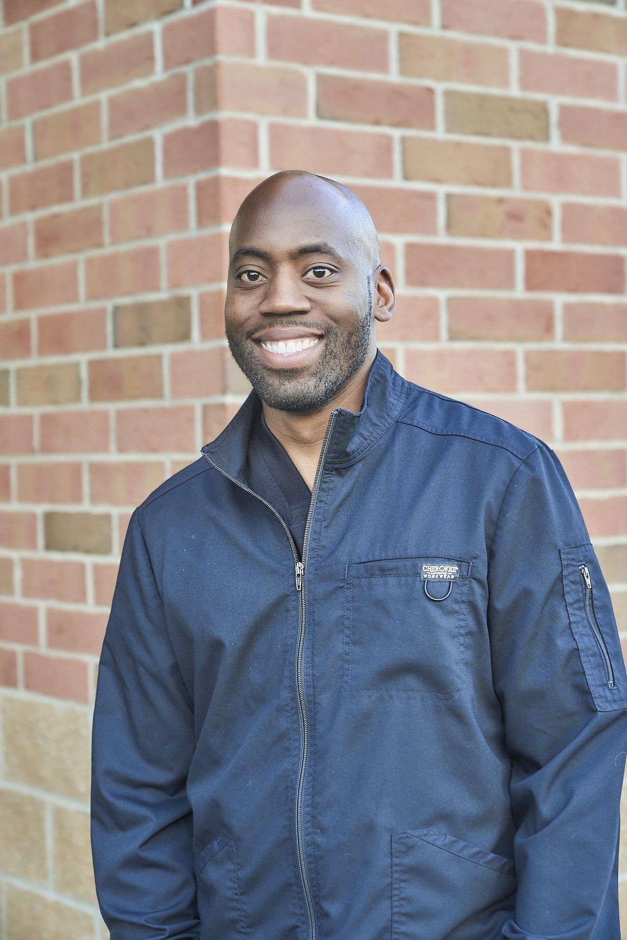 A man in a blue jacket is smiling in front of a brick wall.