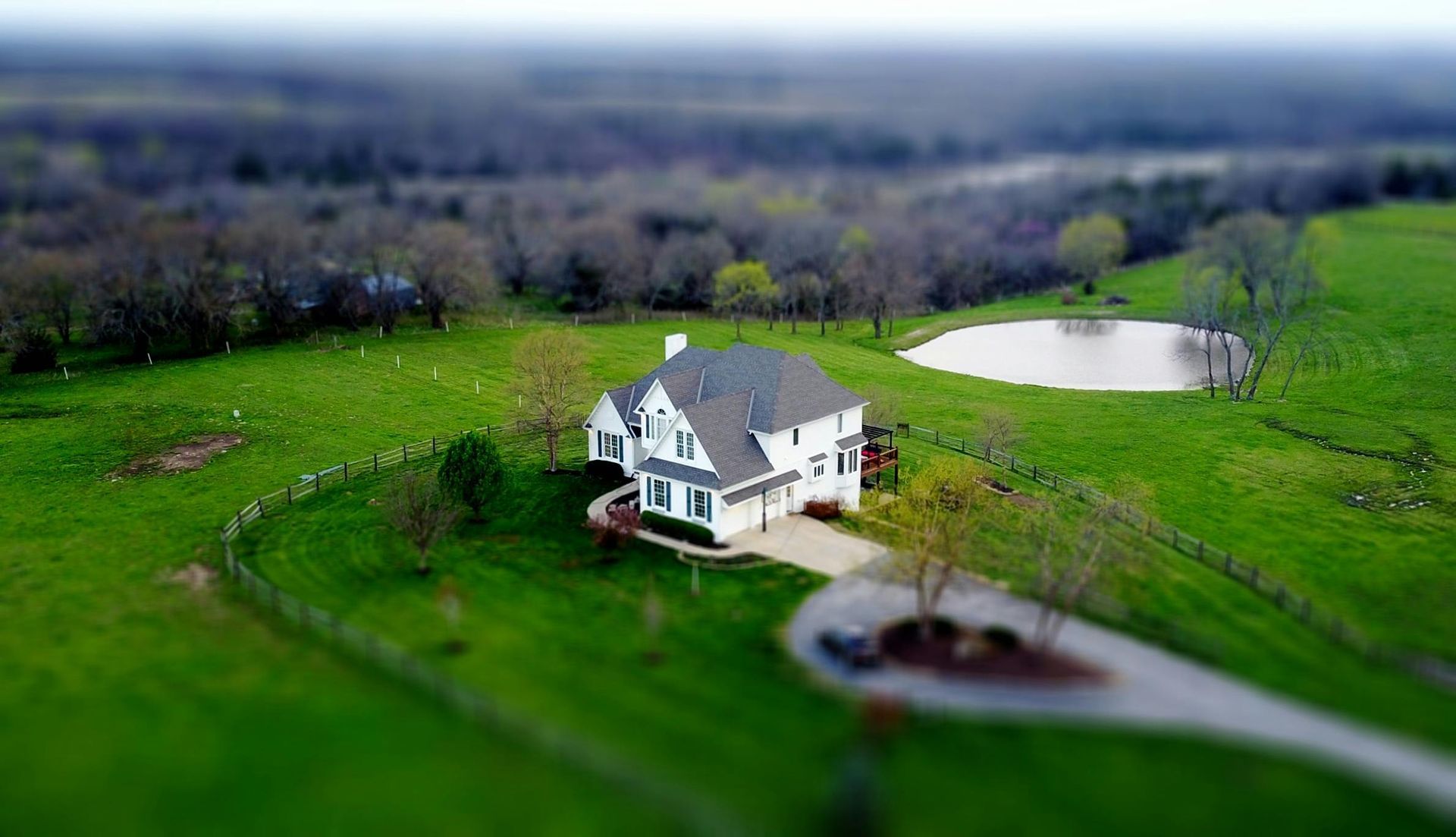 A white house sits in a grassy field next to a small circular pond, viewed from above with a tilt-shift focus effect.