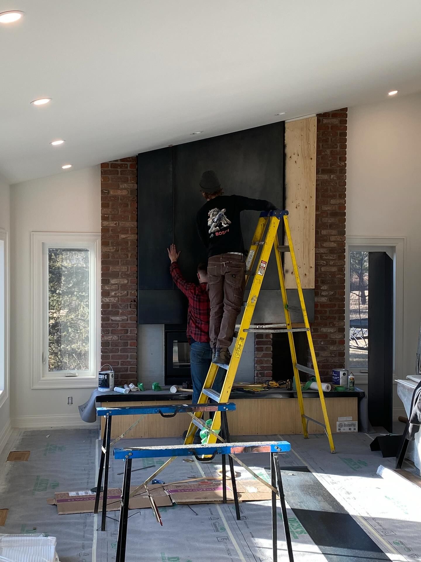 Two people work on a brick fireplace renovation, with one person on a yellow ladder installing large, dark panels.