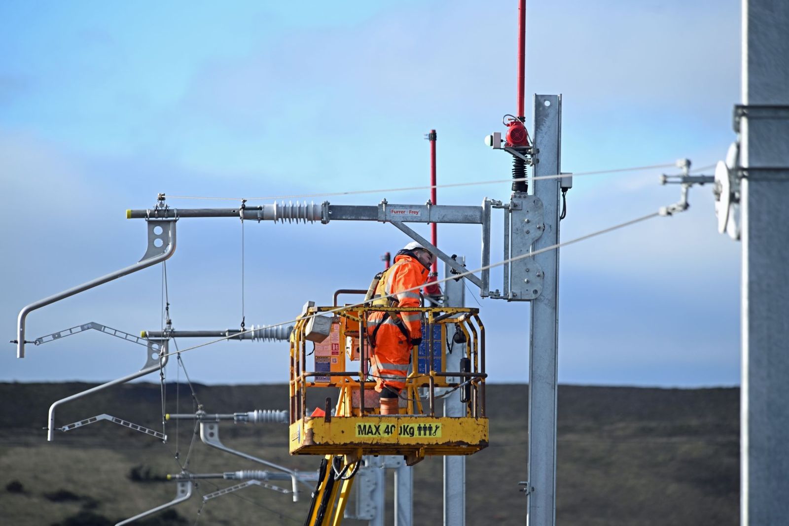 Worker in orange safety gear on a lift working on electrical equipment outdoors.
