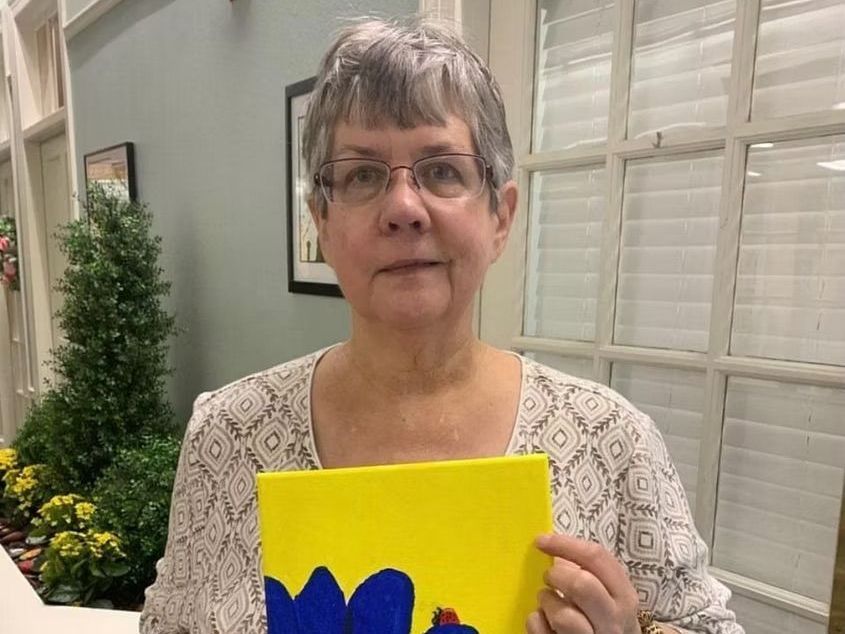 A woman is holding a painting of a blue chicken on a yellow background.