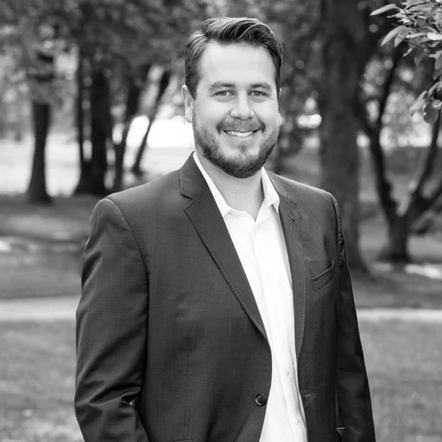 A man in a suit and white shirt is standing in a park.