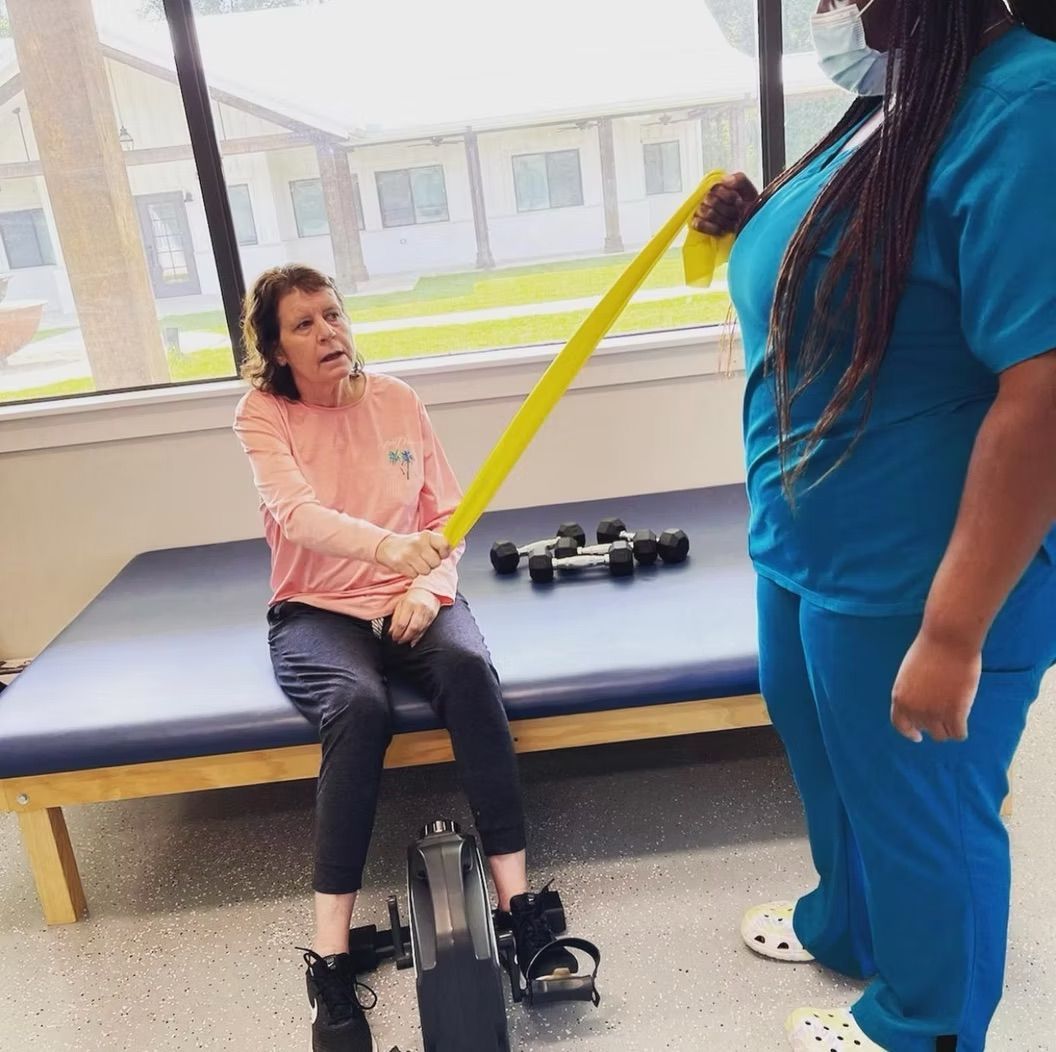 A woman in a pink shirt is sitting on a bed with a yellow resistance band