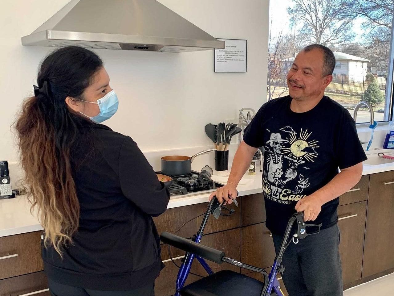 A woman wearing a mask is standing next to a man with a walker in a kitchen.
