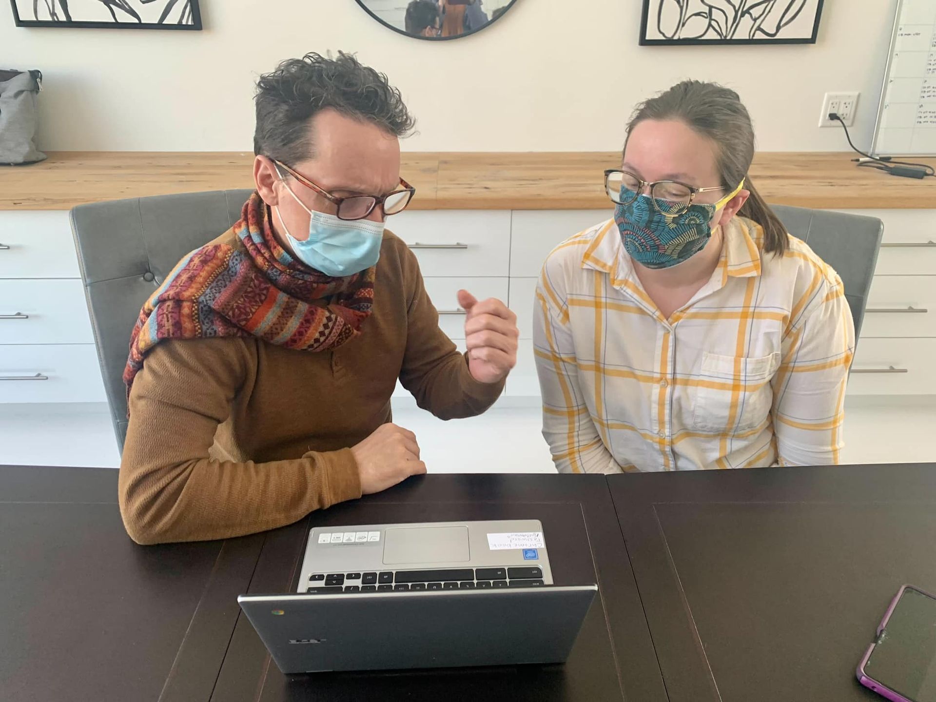 A man and a woman wearing face masks are sitting at a table looking at a laptop.