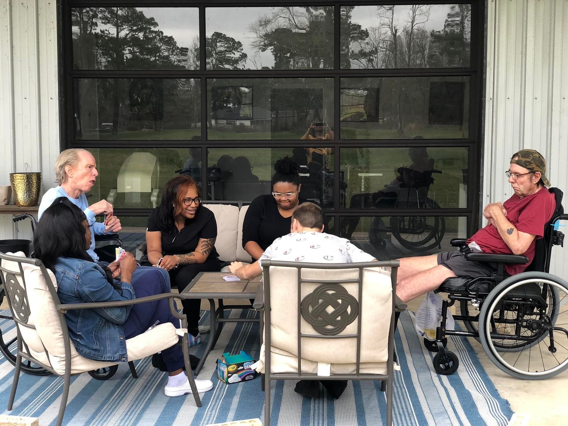 A group of people are sitting around a table on a porch . one of the people is in a wheelchair.