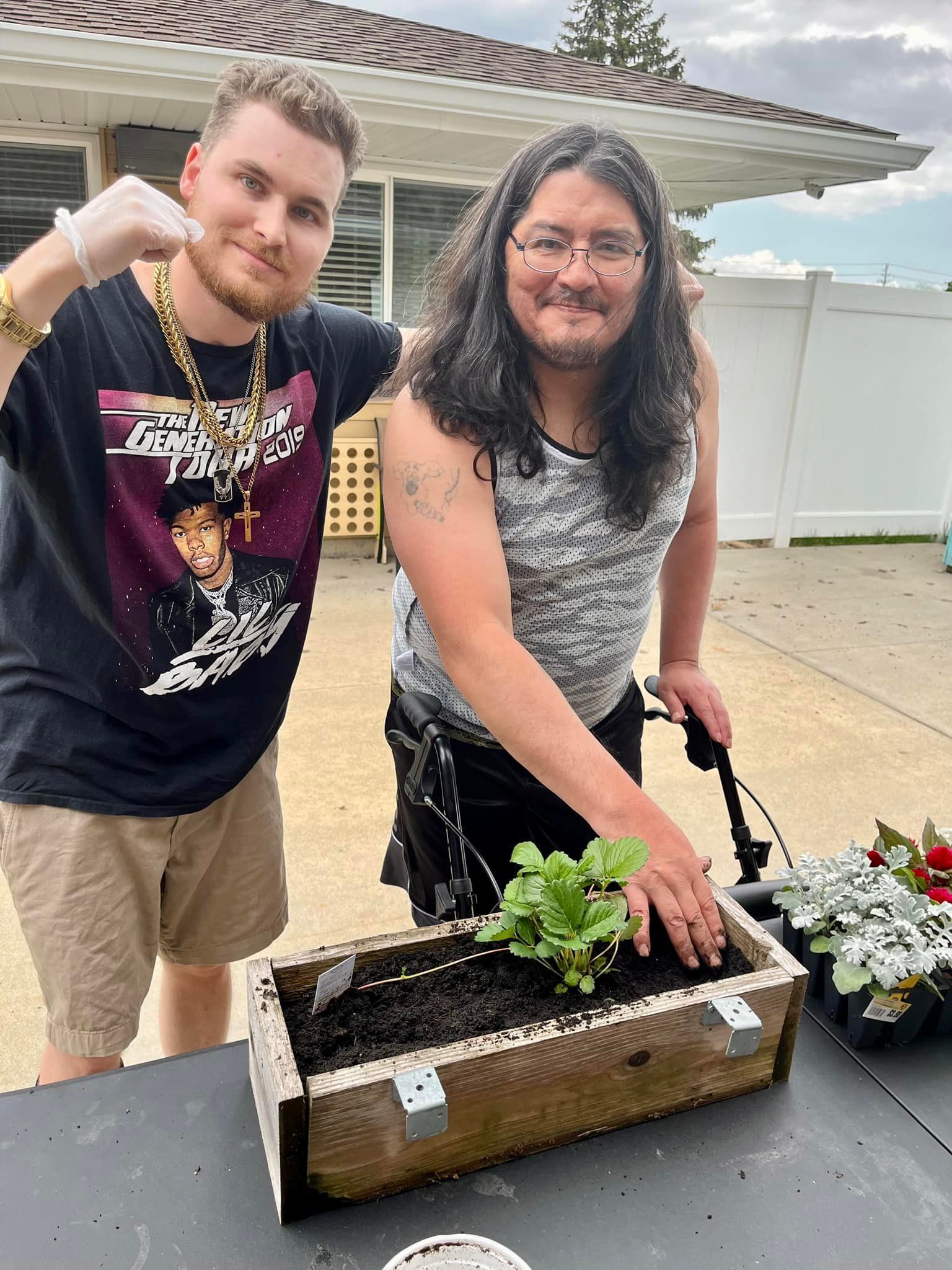 Two men are standing next to each other in front of a wooden box with plants in it.