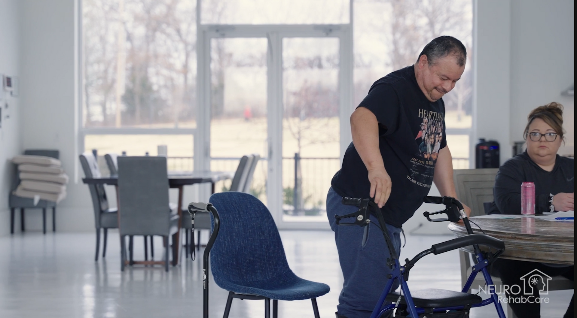A man is using a walker in a room while a woman sits at a table.