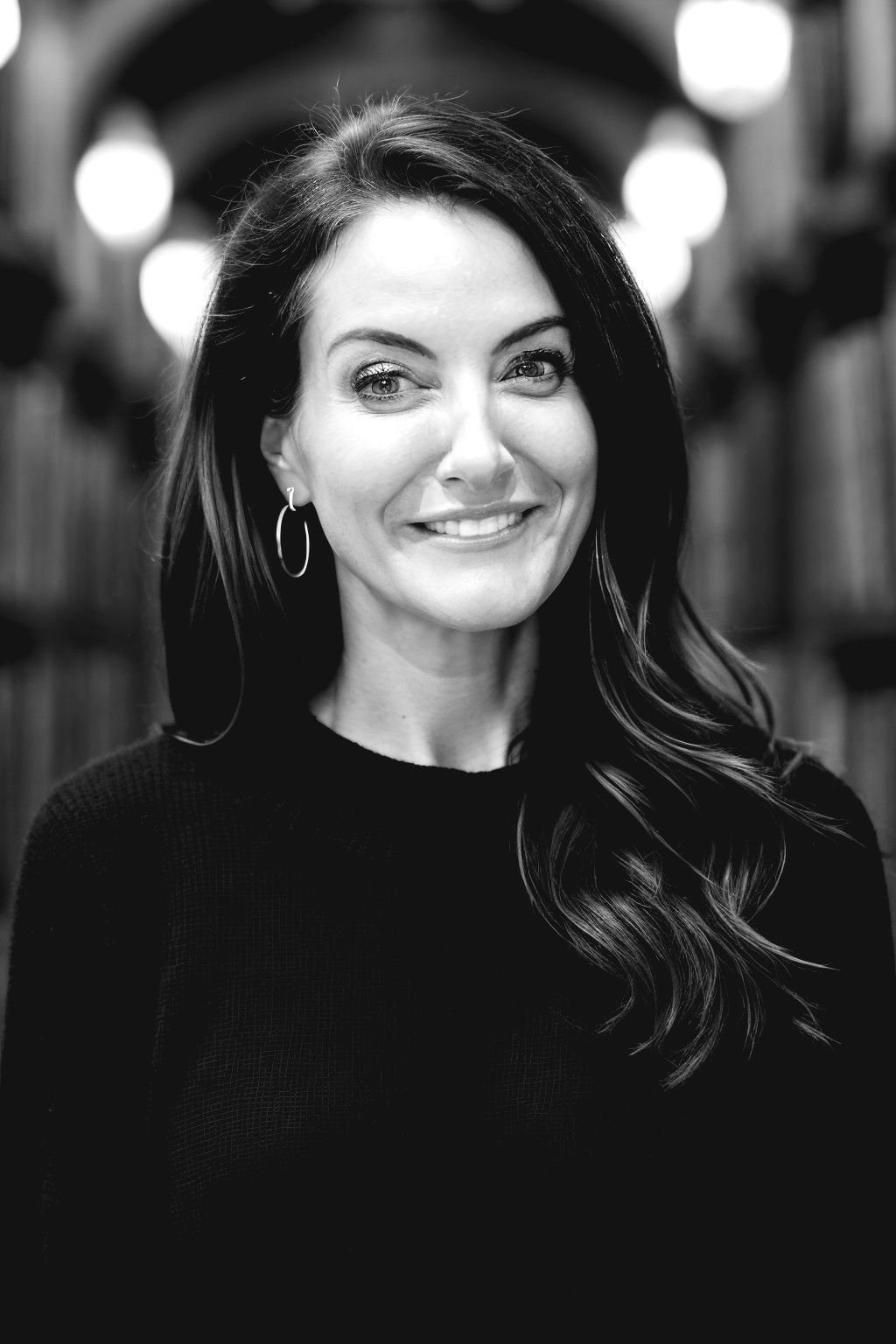 Kristi Husk. A black and white photo of a smiling woman in a library.