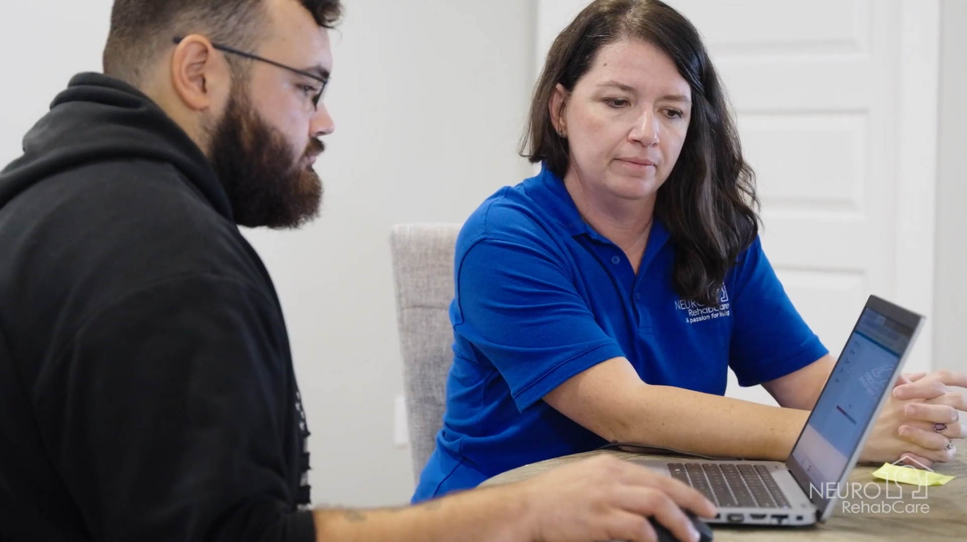 A man and a woman are sitting at a table looking at a laptop computer.