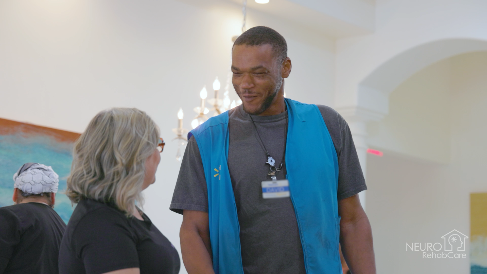 A man in a blue vest is talking to a woman in a room.
