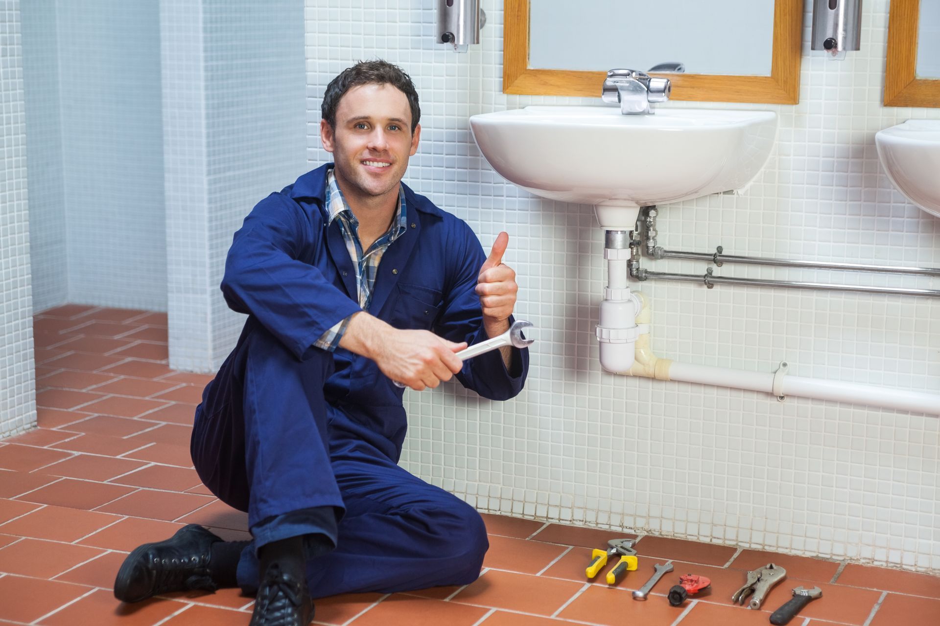 a plumber is kneeling on the floor in a bathroom holding a wrench and giving a thumbs up .