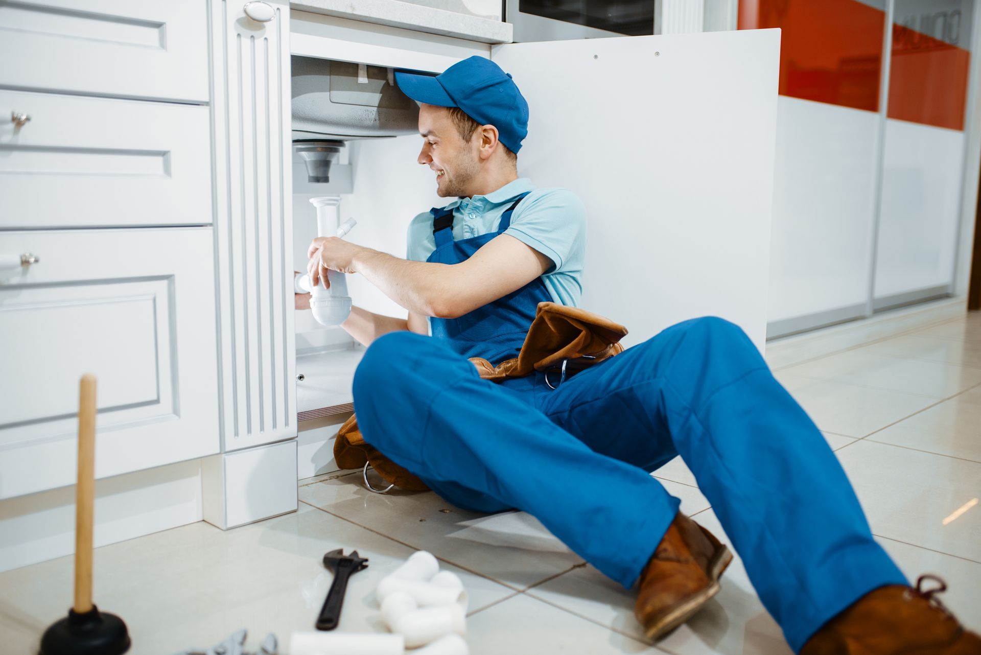 a plumber is sitting on the floor fixing a sink in a kitchen .