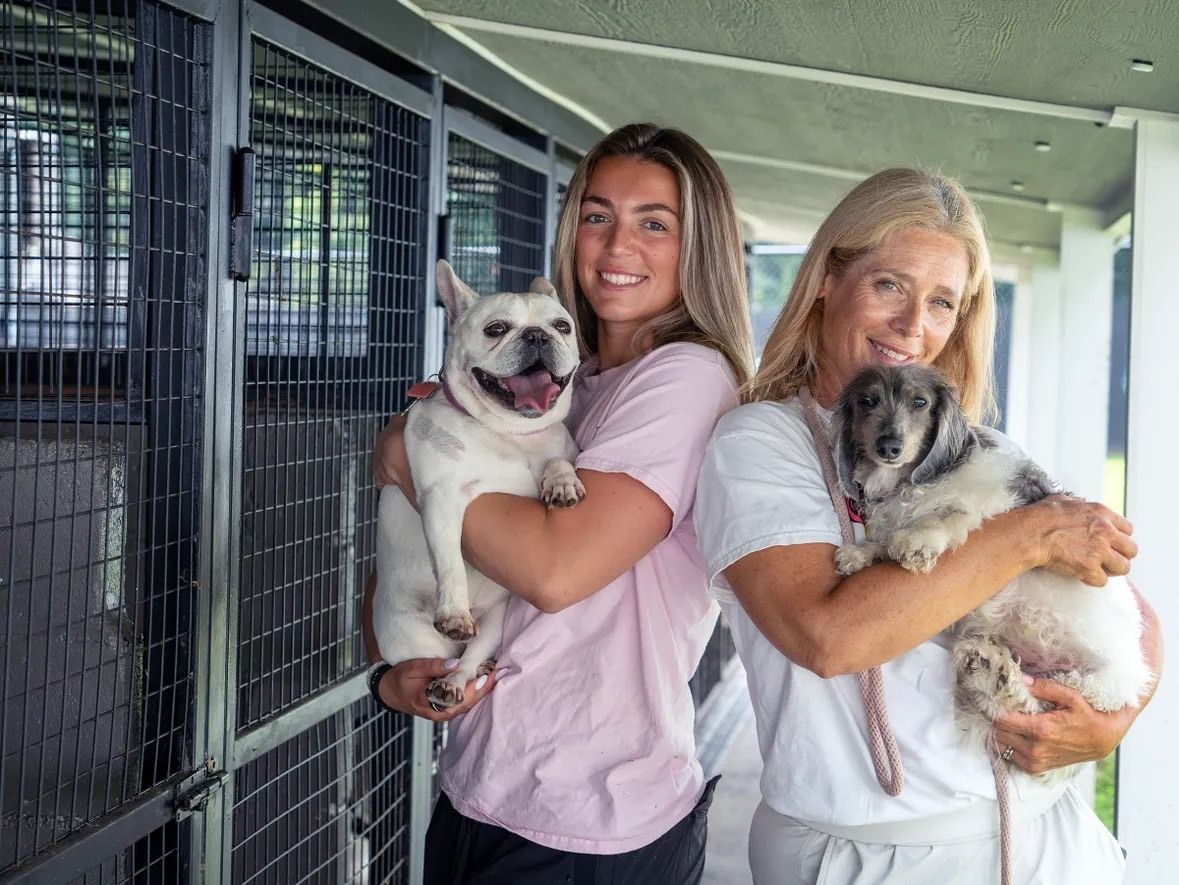 Two people holding dogs in front of outdoor metal kennel enclosures.