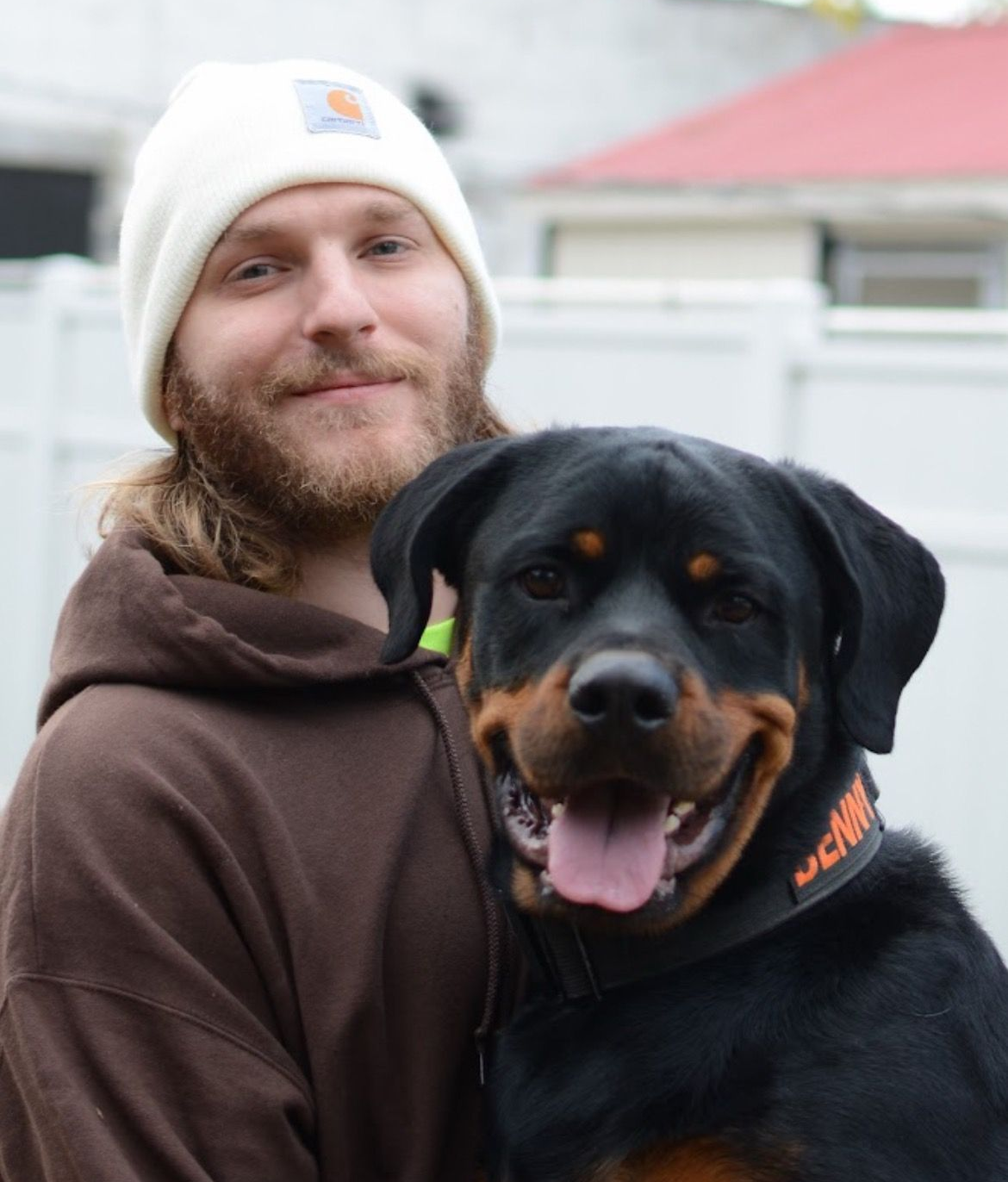 A person in a brown hoodie and white beanie smiles while holding a Rottweiler with an orange collar in a fenced yard.