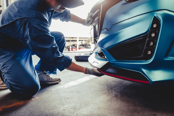 Mechanic in blue jumpsuit inspecting the front bumper of a blue car.