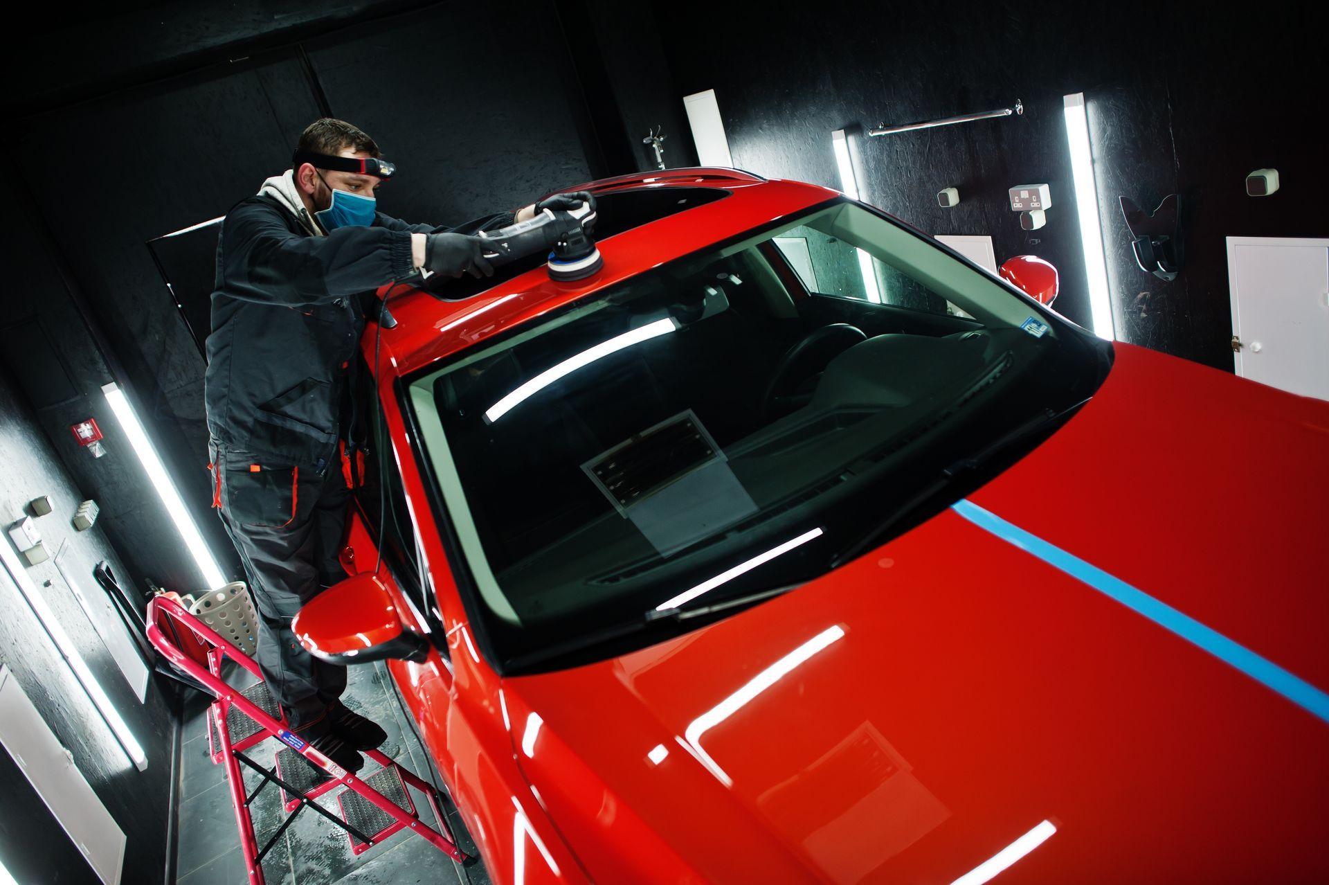 Person polishing a red car's roof with a machine. Blue tape lines roof, setting is a well-lit garage.