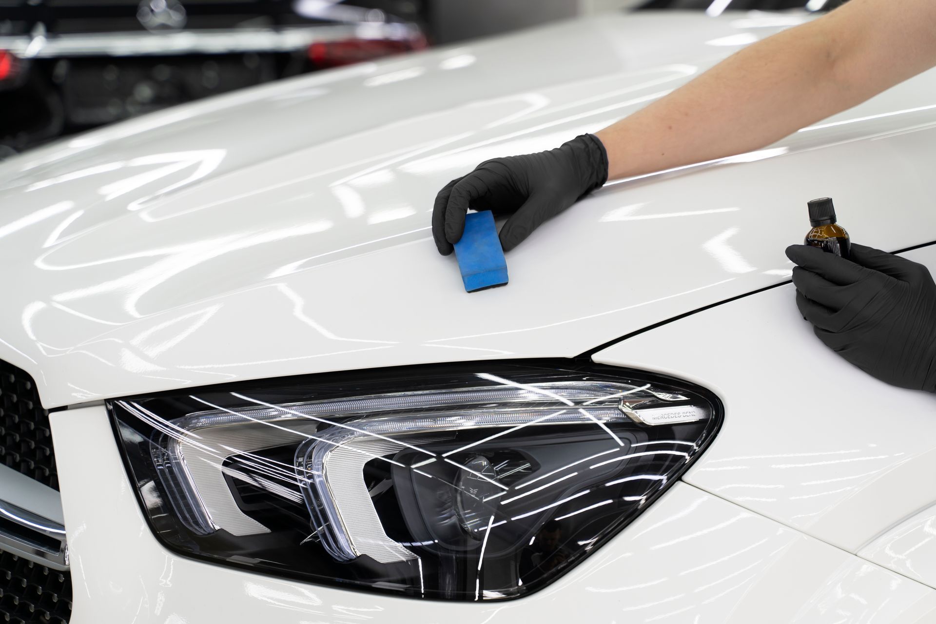 Person applying ceramic coating to a white car hood. Black gloves, blue applicator, and a brown bottle.