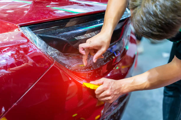 Person applying clear protective film to a red car's bumper using a yellow tool.