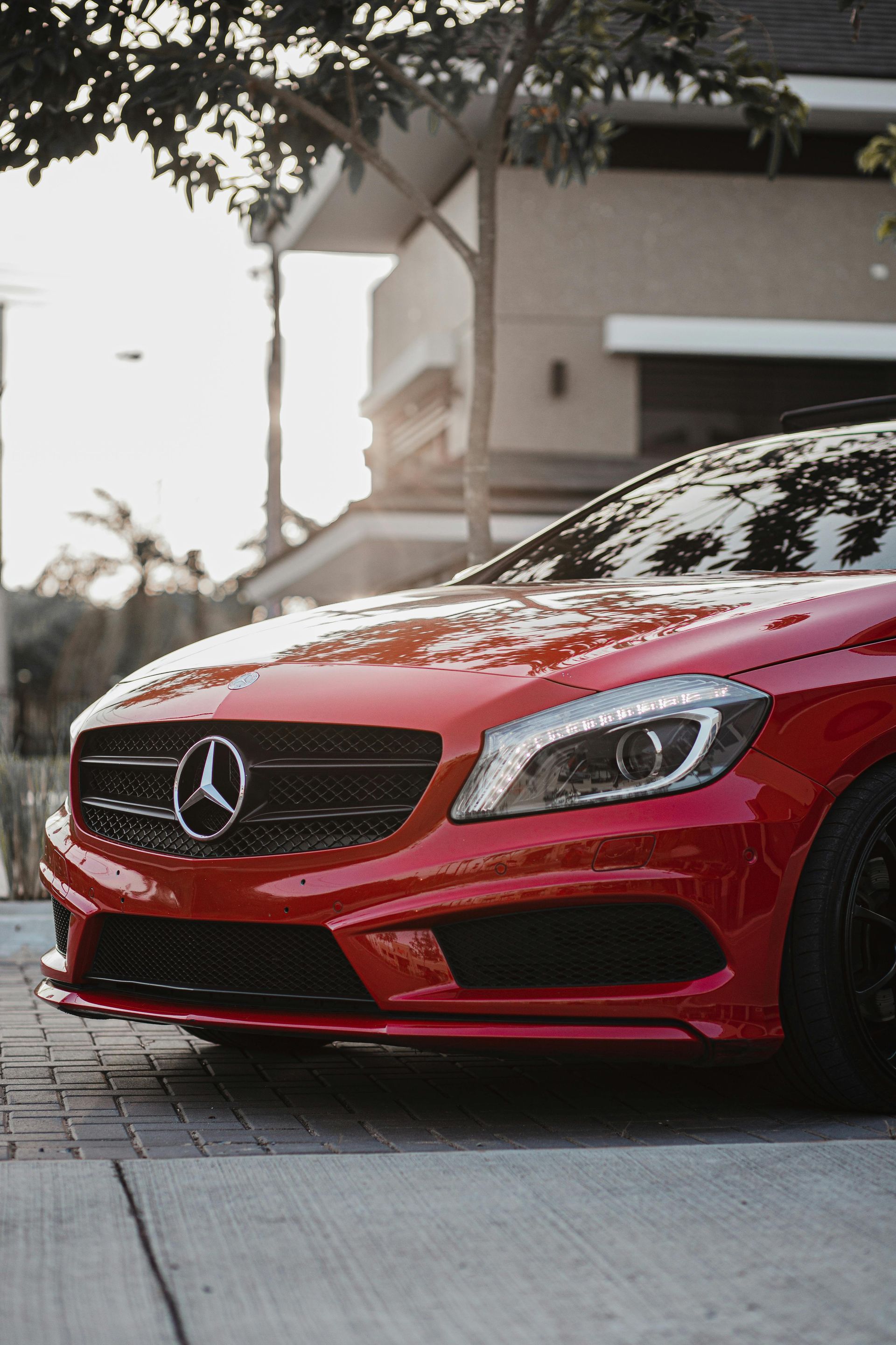 Red Mercedes-Benz car parked on a paved driveway in front of a house, bright sunlight.