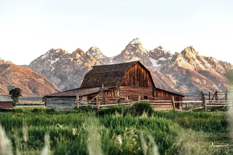 A barn is sitting in the middle of a grassy field with mountains in the background.