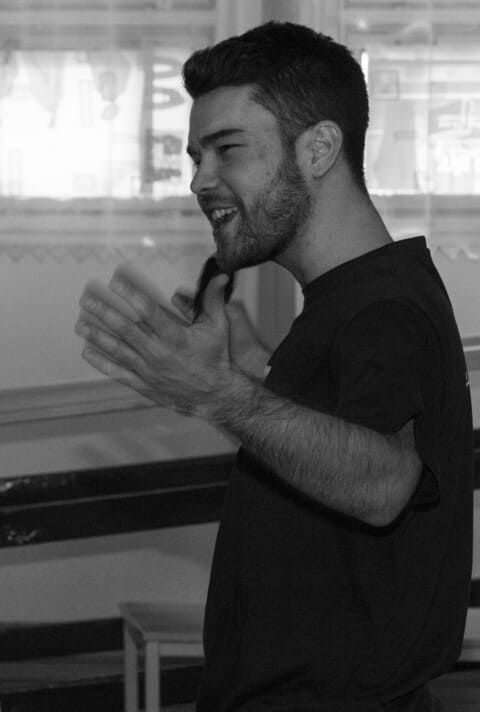 A man with a beard is standing in front of a window with his hands outstretched.