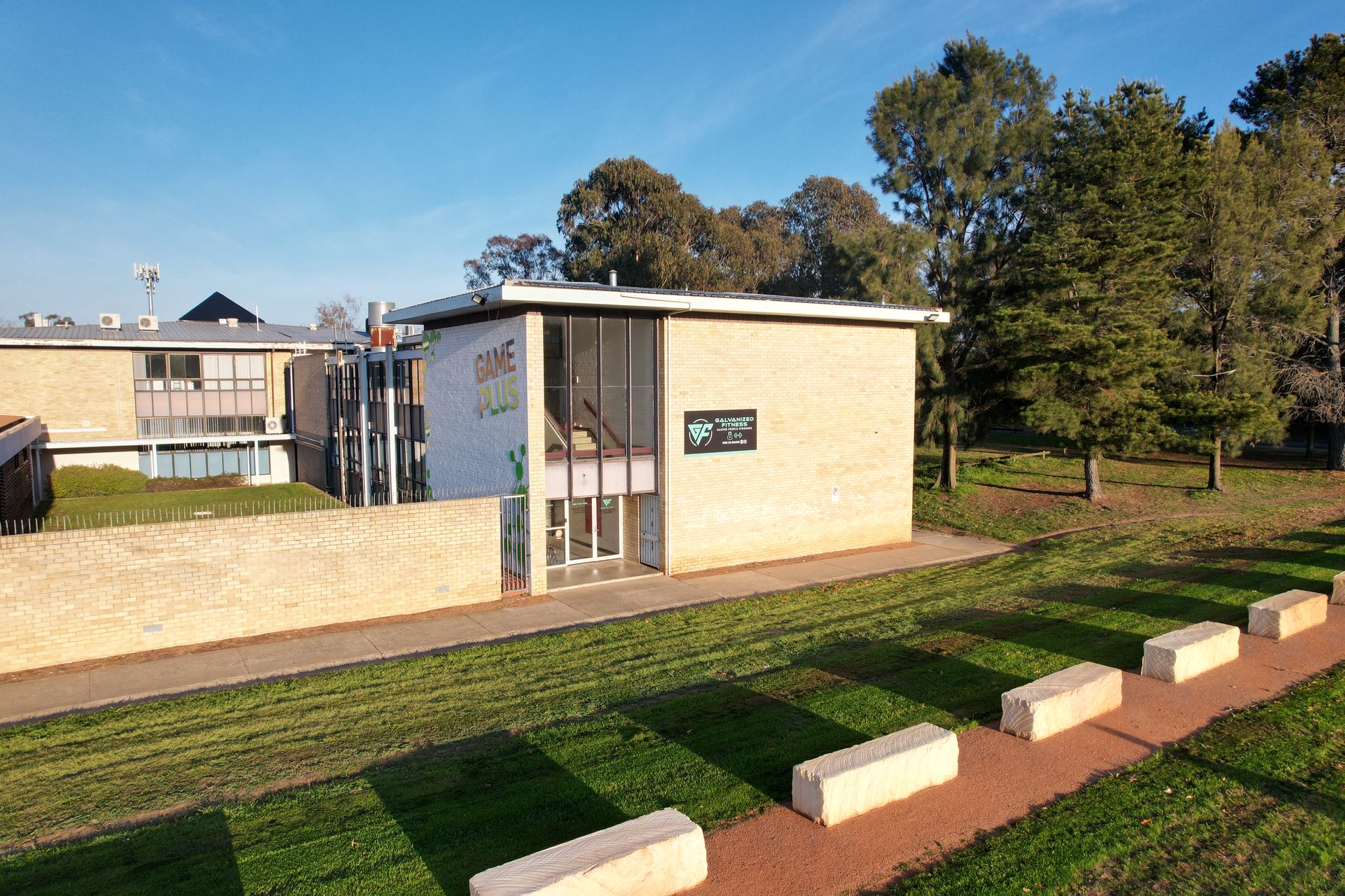 Exterior view of a tan brick building with a glass entrance and a small sign. Sunlight and greenery are present.