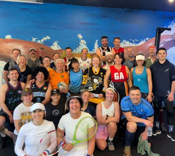 Group photo of diverse people in gym setting, some holding sports equipment and wearing jerseys.