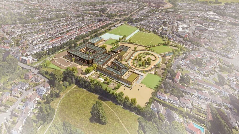 Aerial view of a school complex with multiple buildings, fields, and a circular feature, surrounded by a residential area.