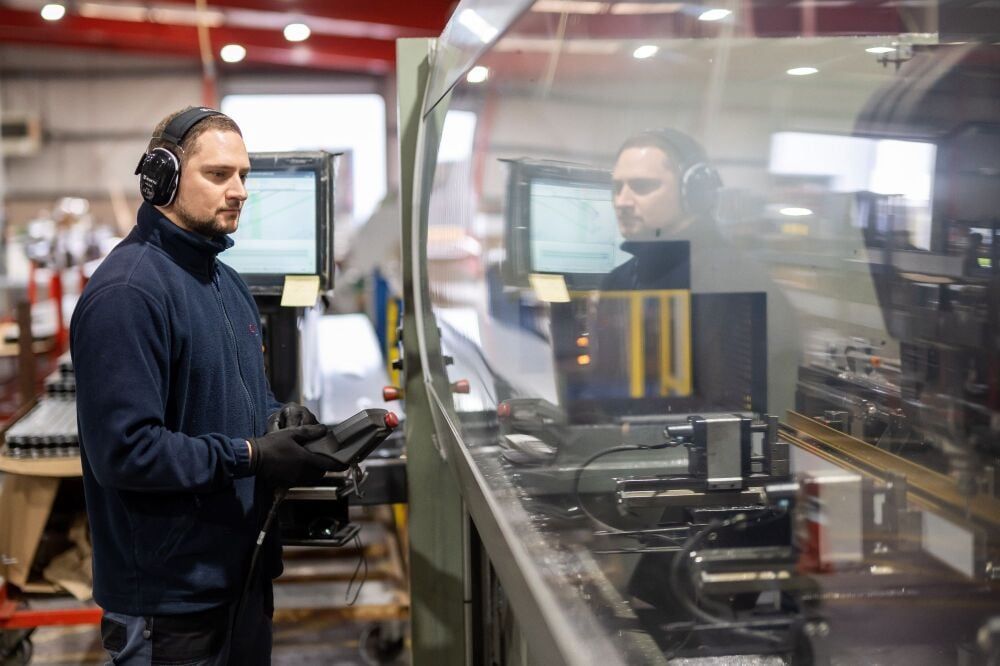 Man in a factory operating machinery, wearing headphones and gloves, focused on a screen.