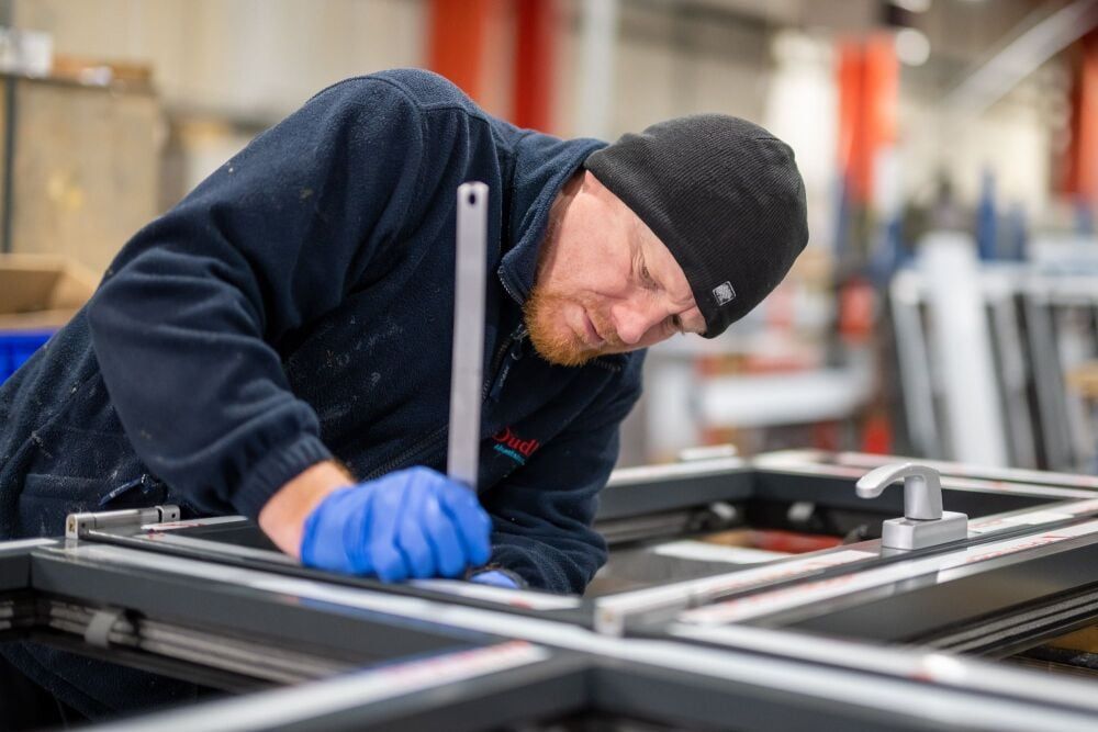 Man in a workshop wearing gloves, beanie, working on a window frame, using a measuring tool.
