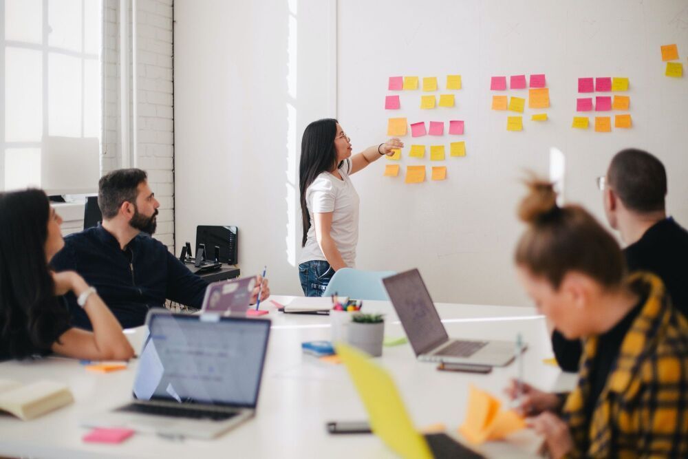 A woman points at sticky notes on a wall during a team meeting.