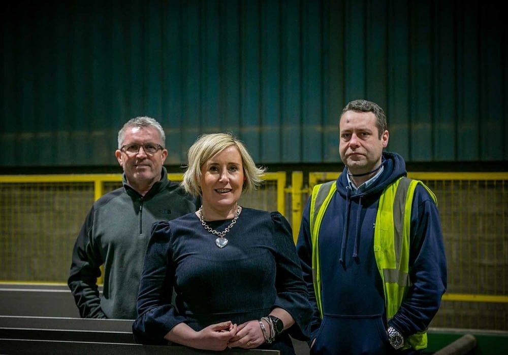 Three people in front of machinery; woman in blue dress, man in gray, and man in safety vest.
