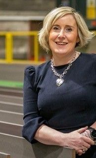 Blonde woman in a navy dress smiles, wearing a silver heart pendant necklace, in a factory setting.