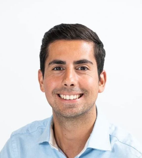 Smiling man with dark hair, wearing a light blue button-down shirt, set against a white background.