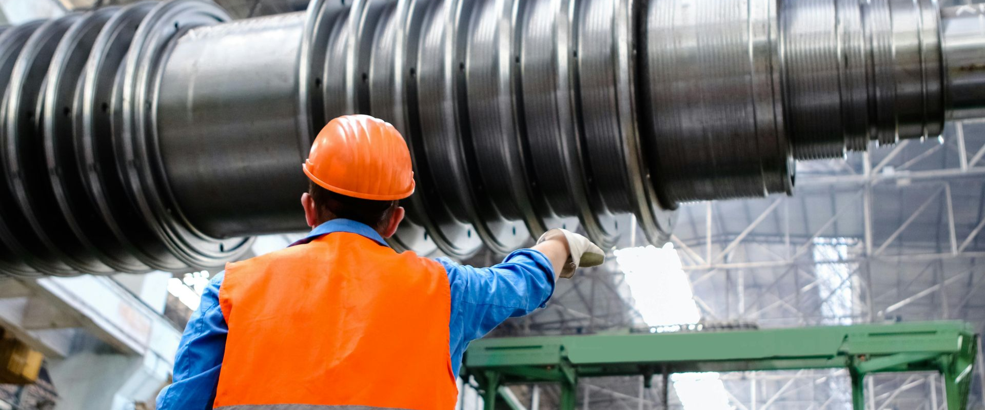 A worker in an orange vest and hard hat examines large industrial machinery, likely in a factory.