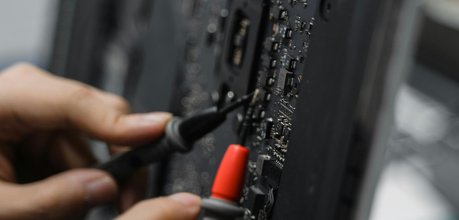 Person tests a circuit board with a multimeter probe. Close-up shows red and black probes on a dark board.